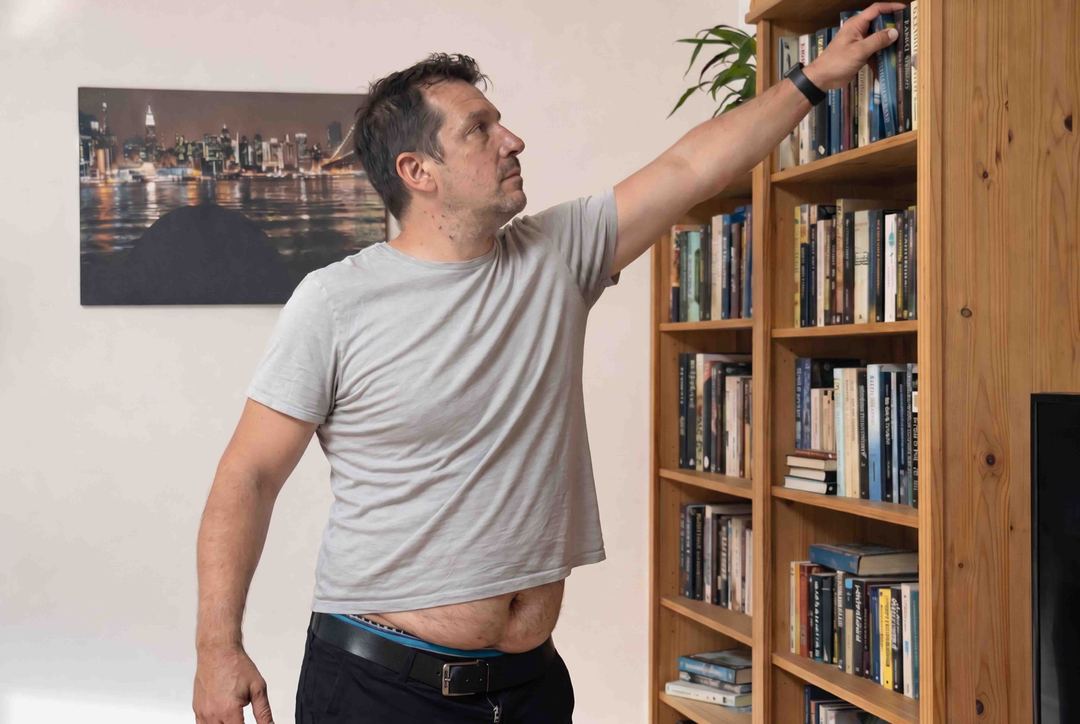 Mann greift nach Bücherregal, das T-Shirt rutscht hoch und gibt den Bauch frei